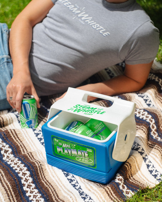 A person lounging on a picnic blanket in the grass holding a can of Steamwhistle pilsner next to a Steamwhistle cooler